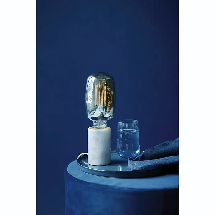 Nordlux Lámpara de sobremesa Siv Mármol - 60 W, L x An x Al: 6 x 6 x 10,3 cm, Blanco, E27 - Una lámpara de mesa elegante con una bombilla Edison y base de cemento, colocada junto a un vaso de agua en un taburete azul.