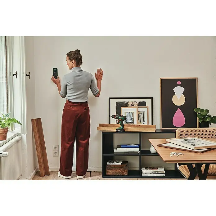 Bosch Ortungsgerät Truvo II - Geeignet für: Aufspüren von spannungsführenden Leitungen und Metall, Erfassungstiefe: Max. 70 mm Eisenmetalle - Eine Frau befestigt mit einem Bosch-Gerät ein Bild an der Wand. Im Hintergrund ist ein Regal mit Bildern und eine Pflanze zu sehen.