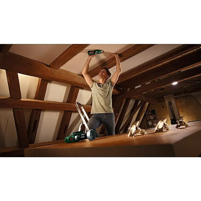 Bosch Ortungsgerät UniversalDetect - Geeignet für: Aufspüren von spannungsführenden Leitungen, Holz und Metall, Erfassungstiefe: Max. 50 mm stromführende Leitung - Mann mit Bosch Ortungsgerät in der Hand, Bohrmaschine und Leuchten auf dem Dachboden.