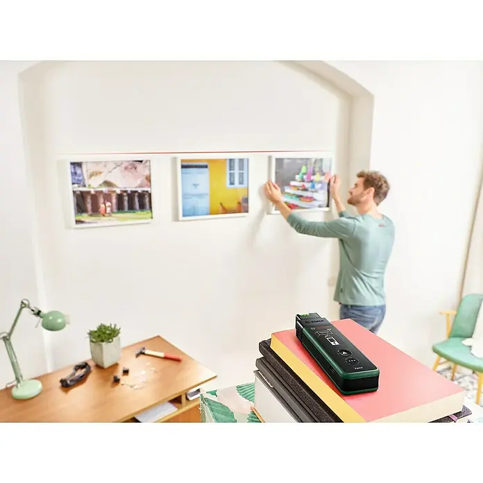 Bosch Linien-Adapter ZamoPassend für: Laser-Entfernungsmesser ZAMO IV - Das Bild zeigt einen Bosch Truvo Leitungssucher auf einem Bücherstapel, während eine Person Bilder aufhängt.