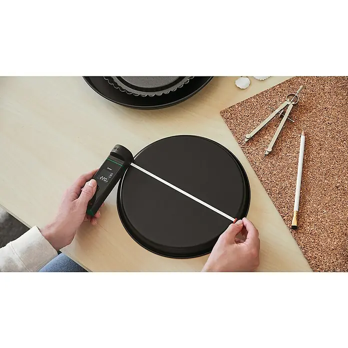 Bosch Bandmaß-Adapter Zamo - Passend für: Laser-Entfernungsmesser ZAMO IV - Eine Person misst den Durchmesser einer schwarzen Kuchenform mit einem Bosch Zamo Laser-Entfernungsmesser auf einem Holztisch.