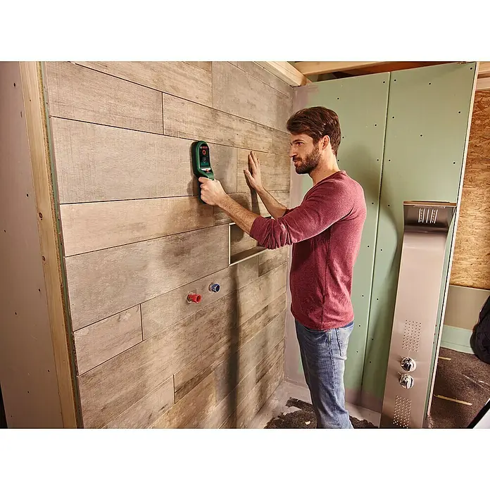 Bosch Ortungsgerät UniversalDetect - Geeignet für: Aufspüren von spannungsführenden Leitungen, Holz und Metall, Erfassungstiefe: Max. 50 mm stromführende Leitung - Ein Mann verwendet einen Bosch-Detektor an einer gefliesten Wand, um Installationen in einem Badezimmer zu überprüfen. Das Gerät hilft, verborgene Leitungen zu finden.