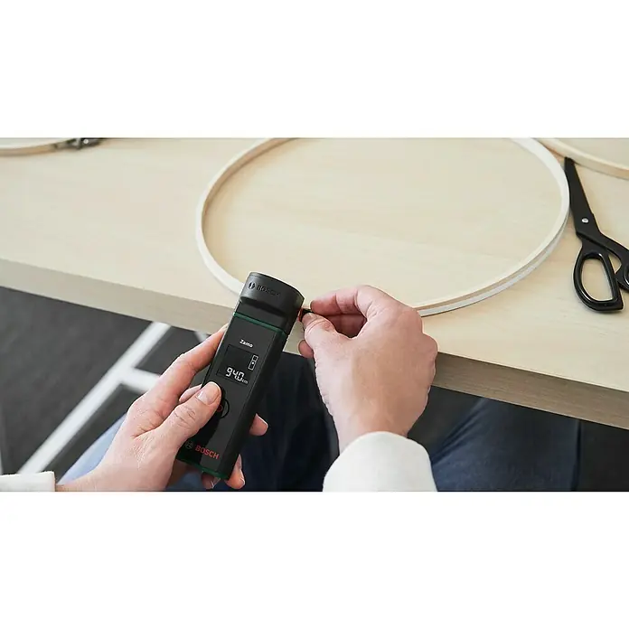 Bosch Bandmaß-Adapter Zamo - Passend für: Laser-Entfernungsmesser ZAMO IV - Ein Bosch Zamo-Lasermessgerät wird verwendet, um ein Holzobjekt zu messen, das auf einem Tisch liegt. Praktisches Werkzeug für genaue Messungen.