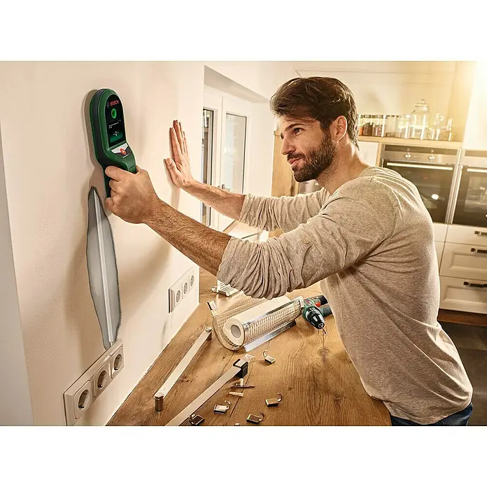Bosch Ortungsgerät UniversalDetect - Geeignet für: Aufspüren von spannungsführenden Leitungen, Holz und Metall, Erfassungstiefe: Max. 50 mm stromführende Leitung - Ein Mann verwendet einen Bosch-Detektor an einer Wand in einer Küche. Das Gerät hilft ihm, verborgene Hindernisse vor der Montage zu finden.