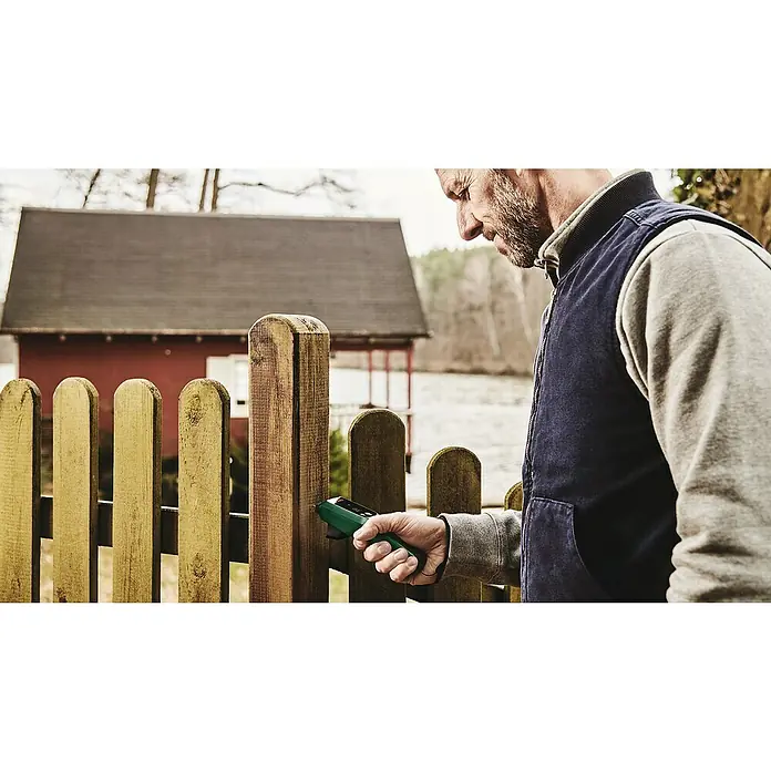 Bosch Universal Feuchtigkeitsmessgerät UniversalHumid - Geeignet für: Holz, Messbereich: 7,1 - 74,7 % - Nahaufnahme eines Mannes, der mit einem Bosch-Feuchtigkeitsmesser die Feuchtigkeit eines Holzzauns misst, mit einem Haus im Hintergrund.