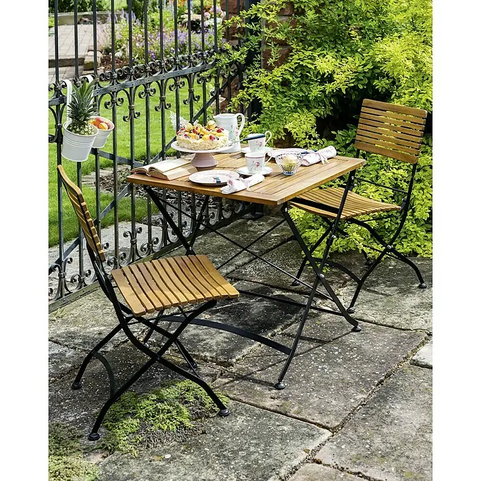Sunfun Moni Balkontisch - L x B x H: 75 x 75 x 75 cm, Akazie, Klappbar, Eckig, Natur/Schwarz - Eine Szene im Freien mit einem Holztisch und Stühlen, dekoriert mit Geschirr und Essen, vor einem Garten mit üppigem Grün.