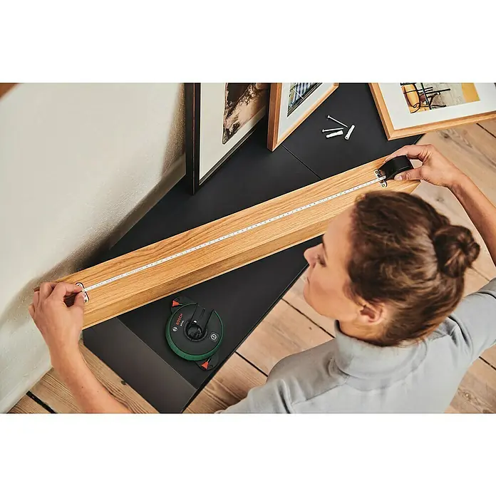 Bosch Linienlaser Atino - Max. Arbeitsbereich: 2 m - 2,2 m - Die Person misst einen Holzrahmen mit einem Rollmaßband, während ein Bosch-Laser-Entfernungsmesser daneben liegt.