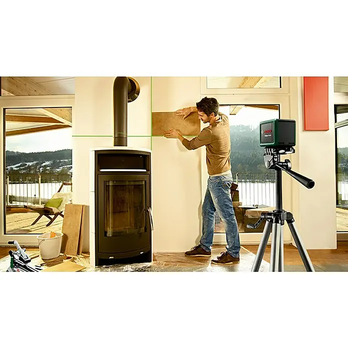 Bosch Kreuzlinienlaser Quigo Green - Max. Arbeitsbereich: 12 m - Ein Mann montiert Fliesen mit Hilfe eines Bosch Linienlasers an einer Wand, der auf einem Stativ steht, neben einem Kamin.