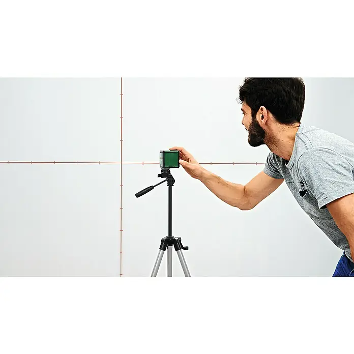 Bosch Kreuzlinienlaser Quigo Plus - Max. Arbeitsbereich: 7 m - Ein Mann justiert einen grünen Bosch Linienlaser auf einem Stativ, der rote Linien auf eine weiße Wand projiziert für präzise Ausrichtung.