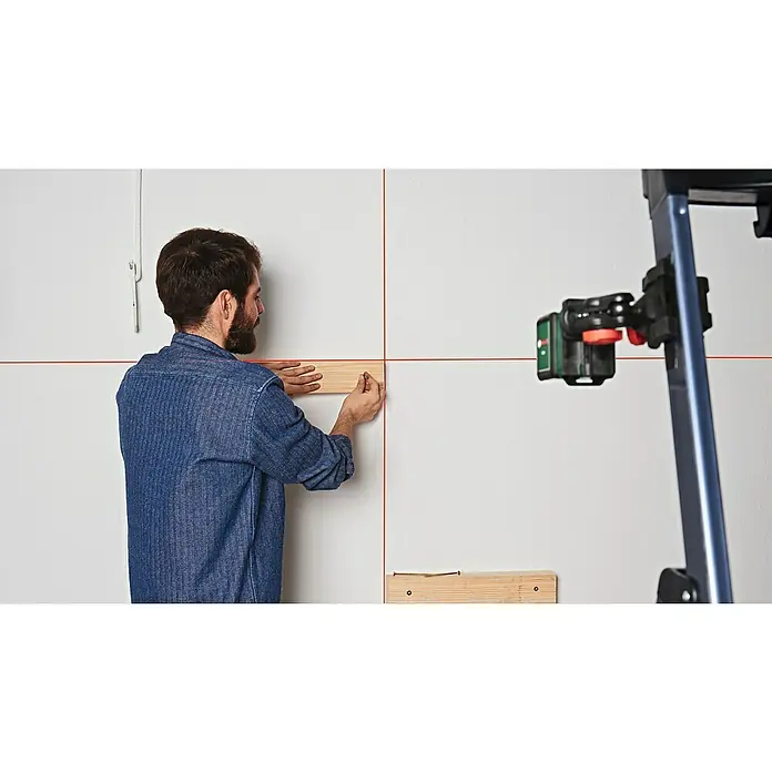 Bosch Kreuzlinienlaser Quigo - Max. Arbeitsbereich: 10 m - Ein Mann mit Bart montiert ein Holzbrett an einer Wand mit Hilfe eines Linienlasers, um die Ausrichtung zu gewährleisten. Das Gerät projiziert rote Linien.