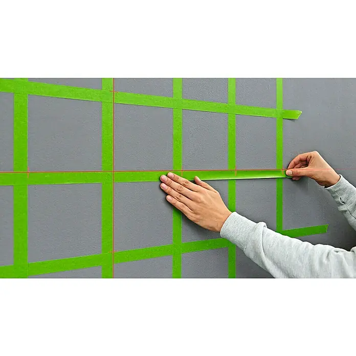 Bosch Kreuzlinienlaser Quigo Plus - Max. Arbeitsbereich: 7 m - Das Bild zeigt, wie grüne Malerkrepp-Bänder mit roter Laserlinie auf einer grauen Wand angebracht werden, um ein geometrisches Muster zu erstellen.