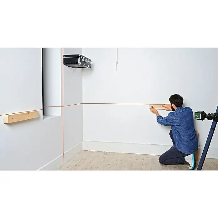 Bosch Kreuzlinienlaser Quigo - Max. Arbeitsbereich: 10 m - Ein Mann kniet vor einer Wand und befestigt ein Holzbrett mit einem Laser, der rote Linien an die Wand projiziert, um die Ausrichtung zu gewährleisten.