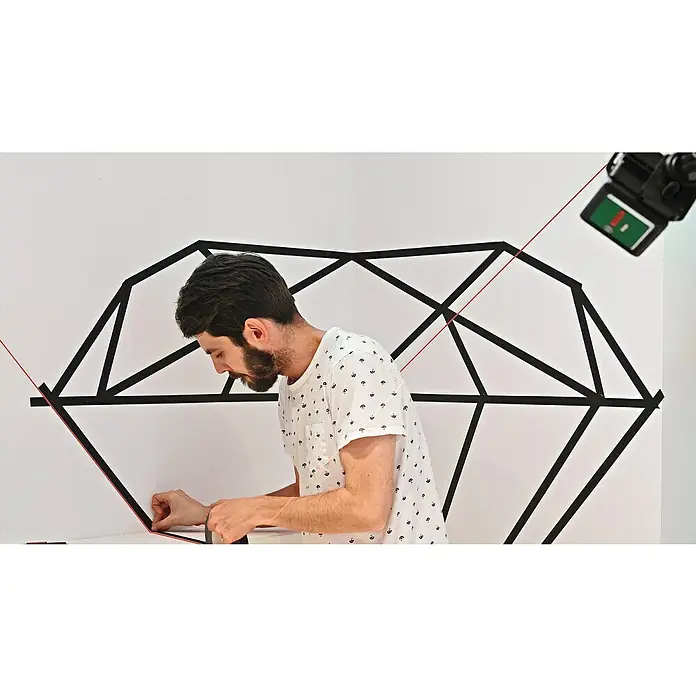Bosch Kreuzlinienlaser Quigo - Max. Arbeitsbereich: 10 m - Mann befestigt mit schwarzem Klebeband ein geometrisches Design an einer weißen Wand, aufgenommen mit einer Kamera.
