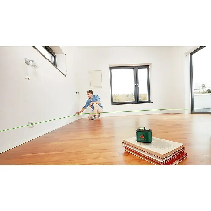 Bosch Kreuzlinienlaser UniversalLevel 360 - Max. Arbeitsbereich: 12 m, 6 -tlg. - Ein grüner Bosch-Laser-Level auf einem Buchstapel mit einer Person, die eine Linie an einer Wand im Zimmer markiert.