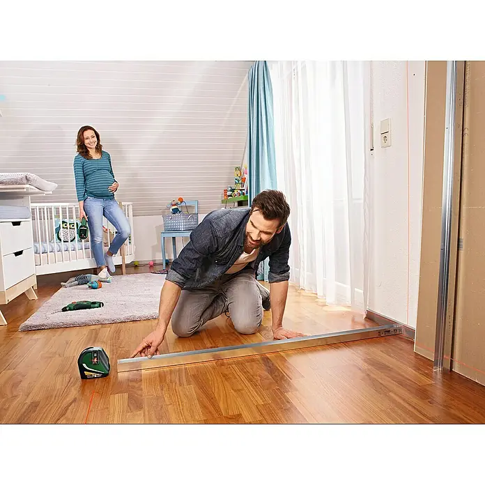 Bosch Kreuzlinienlaser UniversalLevel 3 - Max. Arbeitsbereich: 10 m, 5 -tlg. - Ein Mann kniet auf dem Boden und benutzt einen Bosch-Laser-Entfernungsmesser, um eine gerade Linie zu messen. Eine schwangere Frau steht im Hintergrund.