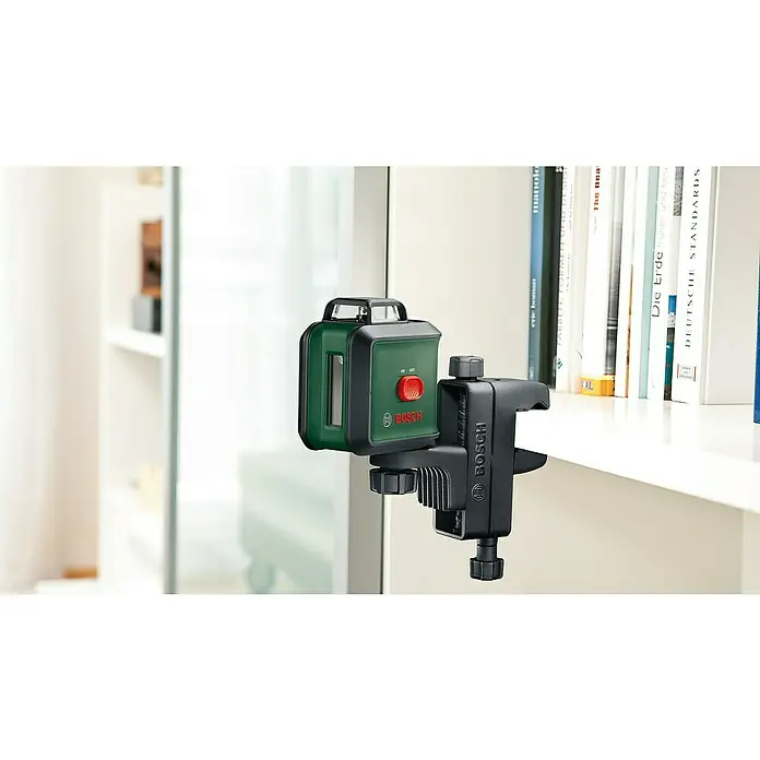 Bosch Klemmhalter MM 3 - Geeignet für: Messgeräte mit ¼″ Gewinde, Arbeitshöhe: 50 cm - 118 cm - Das Bild zeigt einen Bosch Kreuzlinienlaser mit Halterung, befestigt an einer weißen Oberfläche, umgeben von Büchern und Regalen.