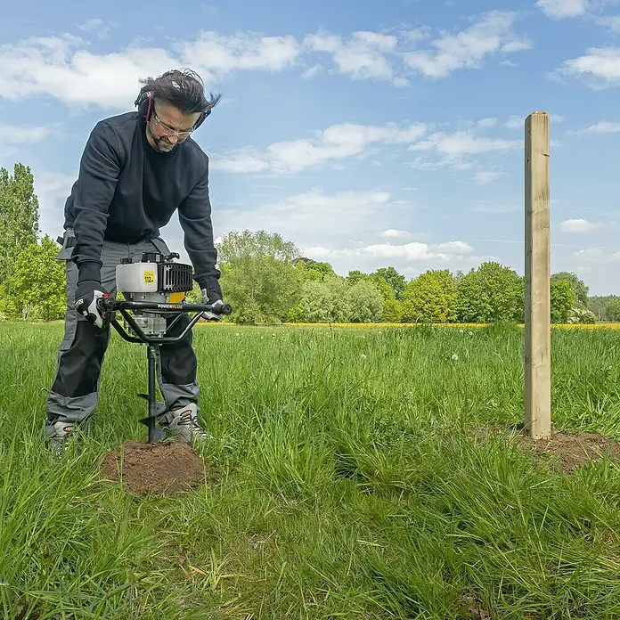 PowerPlus Ahoyadora POWXG50552 - 52 cm³, 1,4 kW, Anchura de trabajo: 10 cm - Un hombre usa una ahoyadora para perforar un agujero para un poste de cerca en un campo verde. La ahoyadora es amarilla y negra.