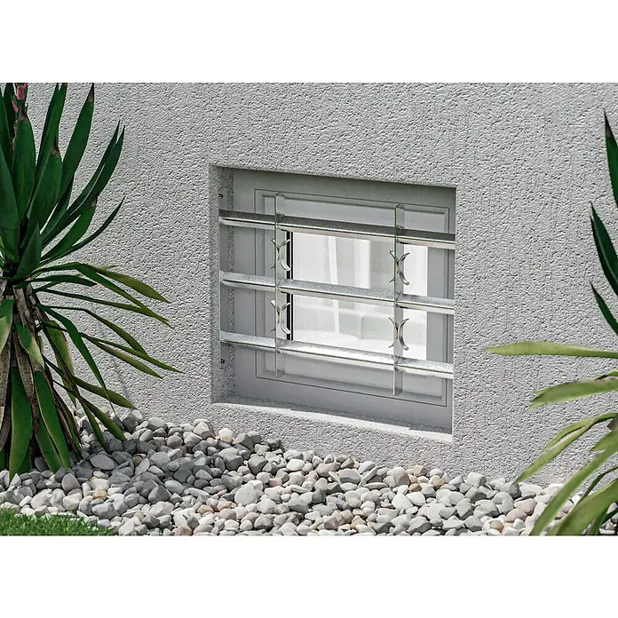 Stabilit Fenstergitter - Höhe: 450 mm, Auszugslänge: 50 cm - 65 cm, Fensterlaibungsmontage - Hier ist ein Kellerfenster mit einem Sicherheitsgitter, umgeben von Kies und Pflanzen. Das Gitter bietet zusätzlichen Schutz für das Fenster.