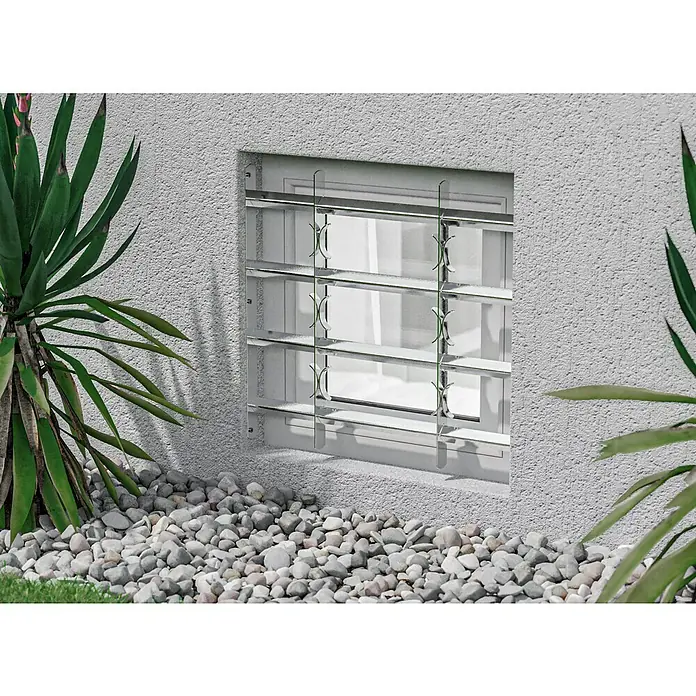 Stabilit Fenstergitter - Höhe: 600 mm, Auszugslänge: 50 cm - 65 cm, Fensterlaibungsmontage - Nahaufnahme eines Fensters mit Sicherheitsgittern aus Metall, umgeben von Kies und Grünpflanzen vor einer strukturierten Wand.