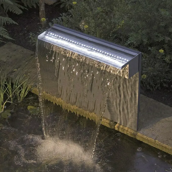 Ubbink Einbau-Wasserfall Niagara LED 60 Set - 12,5 x 60 x 10 cm, Edelstahl, Leuchtmittel: LED - Nahaufnahme eines Wasserfalls mit LED-Beleuchtung, der in einen Teich mündet und von Pflanzen umgeben ist. Der Wasserfall bildet den Mittelpunkt des Gartens.
