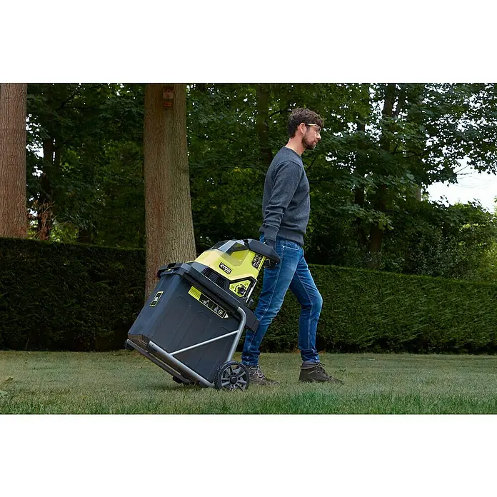 Ryobi Max Power Akku-Häcksler RY36SHX40-0 - Max. Aststärke: 4 cm - Ein Mann zieht einen RYOBI-Gartenhäcksler auf einem Rasen. Der Häcksler ist gelb und schwarz und steht auf einem silbernen Rahmen mit Rädern.