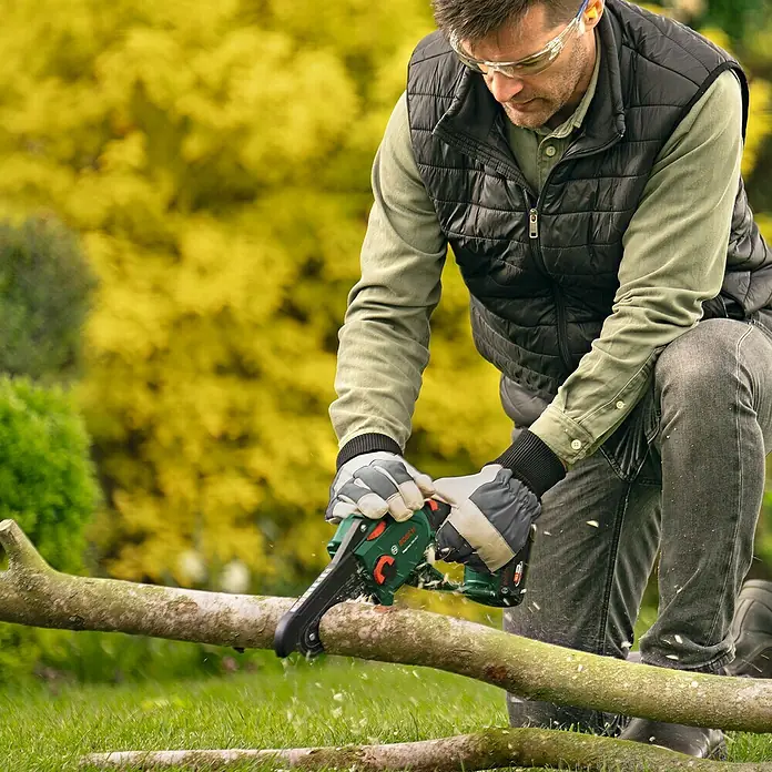 Bosch Power for All 18V Akku-Kettensäge EasyChain 18V-15-7 - 18 V, Li-Ionen, 1 Akku, Schwertlänge: 15 cm - Ein Mann kniet im Garten und sägt mit einer Bosch Akku-Kettensäge einen Ast. Er trägt Schutzbrille und Handschuhe.