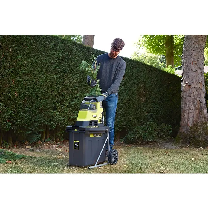 Ryobi Max Power Akku-Häcksler RY36SHX40-0 - Max. Aststärke: 4 cm - Ein Mann benutzt einen Ryobi Häcksler im Garten, um Gartenabfälle zu zerkleinern. Der Häcksler steht vor einer grünen Hecke.