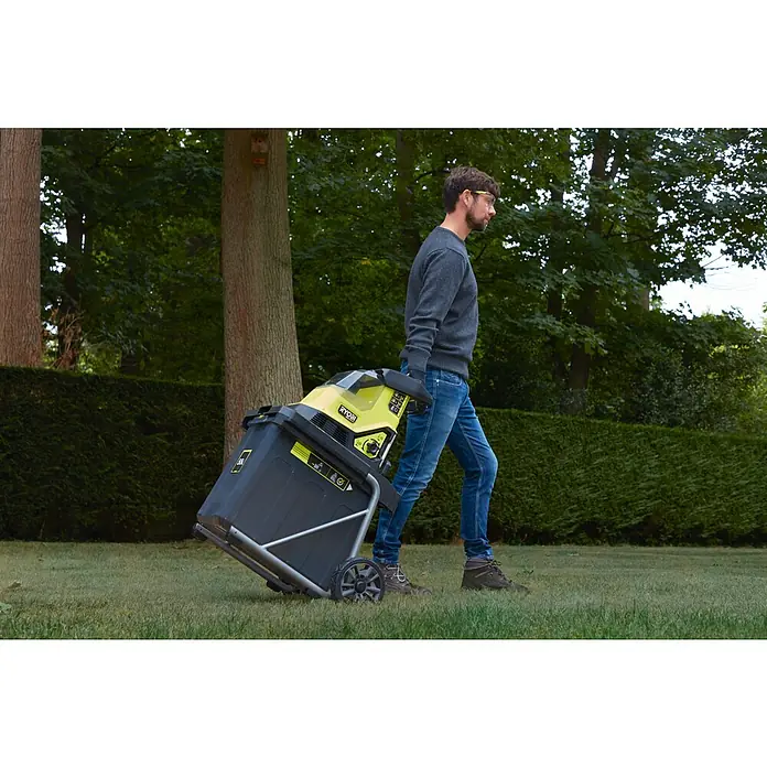 Ryobi Max Power Akku-Häcksler RY36SHX40-0 - Max. Aststärke: 4 cm - Ein Mann zieht einen Ryobi Laubhäcksler mit Fangkorb über einen Rasen vor üppigem Grün. Das Gerät ist gelb und schwarz.
