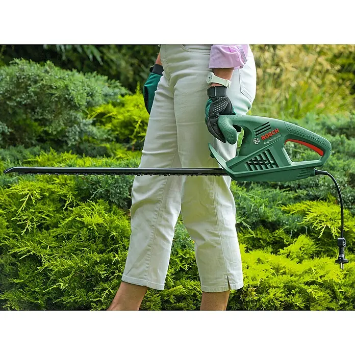 Bosch Elektro-Heckenschere EasyHedgeCut 60 - 450 W, Schnittlänge: 60 cm - Eine Person hält eine grüne Bosch EasyHedgeCut 60 Heckenschere vor einem üppigen grünen Hintergrund.