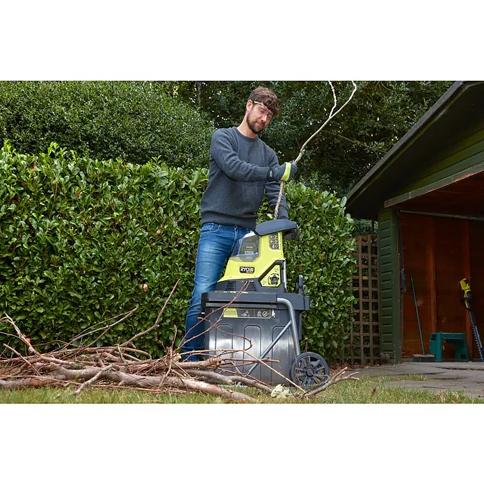 Ryobi Max Power Akku-Häcksler RY36SHX40-0 - Max. Aststärke: 4 cm - Ein Mann benutzt einen RYOBI Häcksler im Garten, um Äste zu zerkleinern, mit einer grünen Hecke und einem Schuppen im Hintergrund.