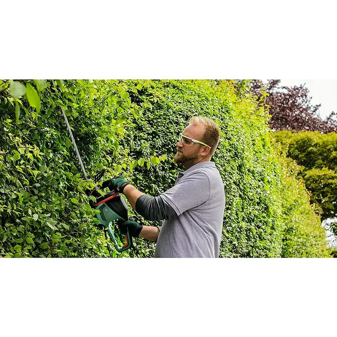 Bosch Elektro-Heckenschere EasyHedgeCut 60 - 450 W, Schnittlänge: 60 cm - Ein Mann, der eine grüne Bosch-Heckenschere zum Trimmen einer dichten Hecke im Garten verwendet. Schutzkleidung wird auch getragen.