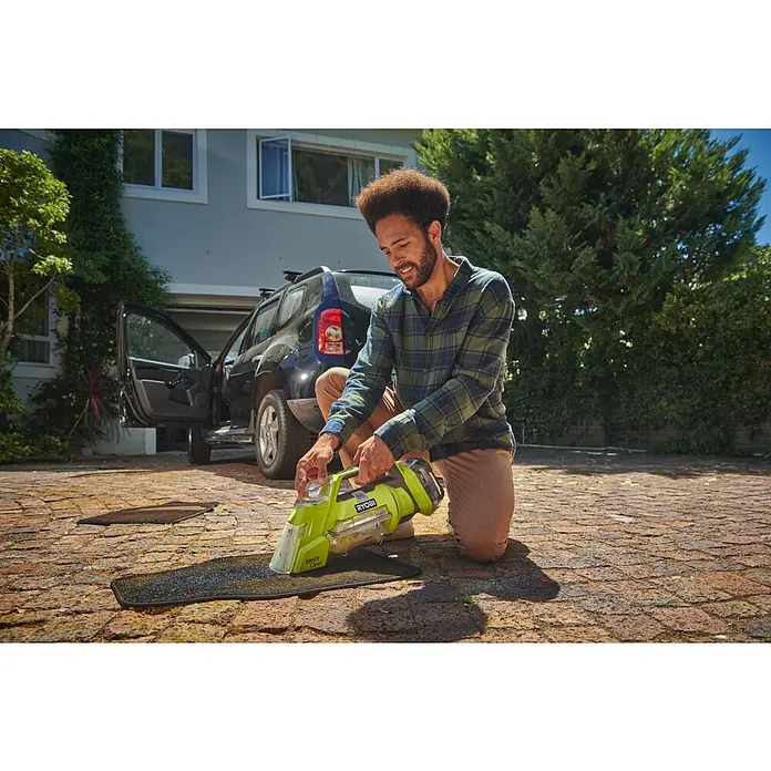 Ryobi ONE+ Akku-Nass-Trockensauger RDC18-0 - 18 V, Ohne Akku, Behältervolumen: 200 l - Ein Mann reinigt mit dem RYOBI Swift Light Spot Cleaner eine schwarze Fußmatte vor einem Auto in der Auffahrt.