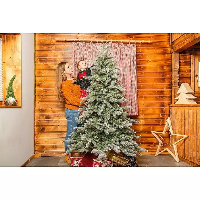 Künstlicher Weihnachtsbaum Frost - Höhe: 180 cm, Grün, Standfuß - Mutter und Kind lächeln neben einem schneebedeckten Weihnachtsbaum mit Geschenken in einem festlichen Holzraum.