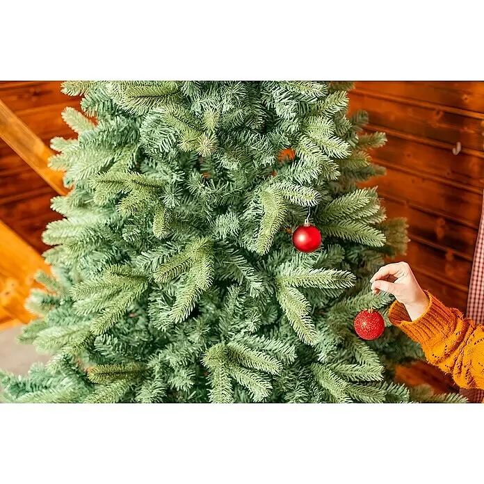 50 NRTH Künstlicher Weihnachtsbaum Vermont - Höhe: 180 cm, Grün, Standfuß - Eine Hand in einem orangen Pullover hängt eine glitzernde rote Christbaumkugel an einen grünen Weihnachtsbaum.