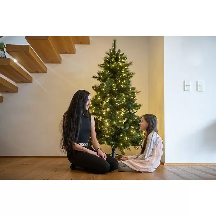 Künstlicher Weihnachtsbaum Mesa - Höhe: 210 cm, Grün, LED-Beleuchtung - Eine Frau und ein Mädchen sitzen auf einem Holzboden und schauen sich neben einem vorbeleuchteten Weihnachtsbaum mit warmen Lichtern an.