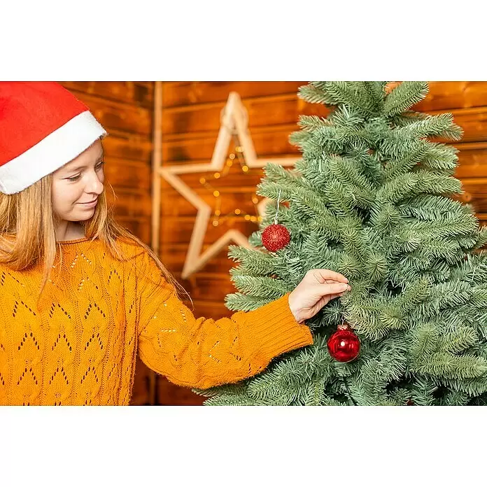 50 NRTH Künstlicher Weihnachtsbaum Vermont - Höhe: 180 cm, Grün, Standfuß - Eine Person in einem orangefarbenen Pullover und einer Weihnachtsmütze hängt eine rote Kugel an einen Weihnachtsbaum.
