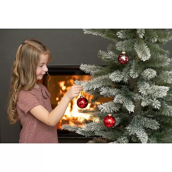 Künstlicher Weihnachtsbaum Cedar Frost - Höhe: 210 cm, Grün, Standfuß - Ein junges Mädchen hängt eine rote Glaskugel an einen beschneiten Weihnachtsbaum.