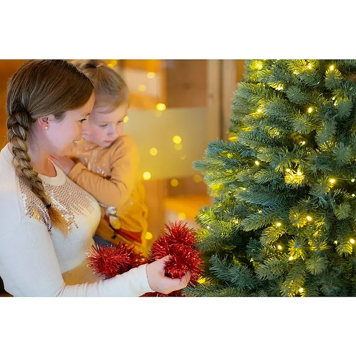 50 NRTH Künstlicher Weihnachtsbaum Vermont - Höhe: 180 cm, Grün, LED-Beleuchtung - Eine Mutter schmückt mit ihrem Kind einen leuchtenden Weihnachtsbaum mit roter Lametta-Girlande, um eine festliche und herzliche Weihnachtsatmosphäre zu schaffen.