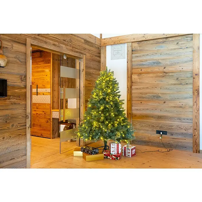 50 NRTH Künstlicher Weihnachtsbaum Vermont - Höhe: 180 cm, Grün, LED-Beleuchtung - Ein festlich geschmückter Weihnachtsbaum mit warmen Lichtern und liebevoll verpackten Geschenken in einem gemütlichen, rustikalen Raum.
