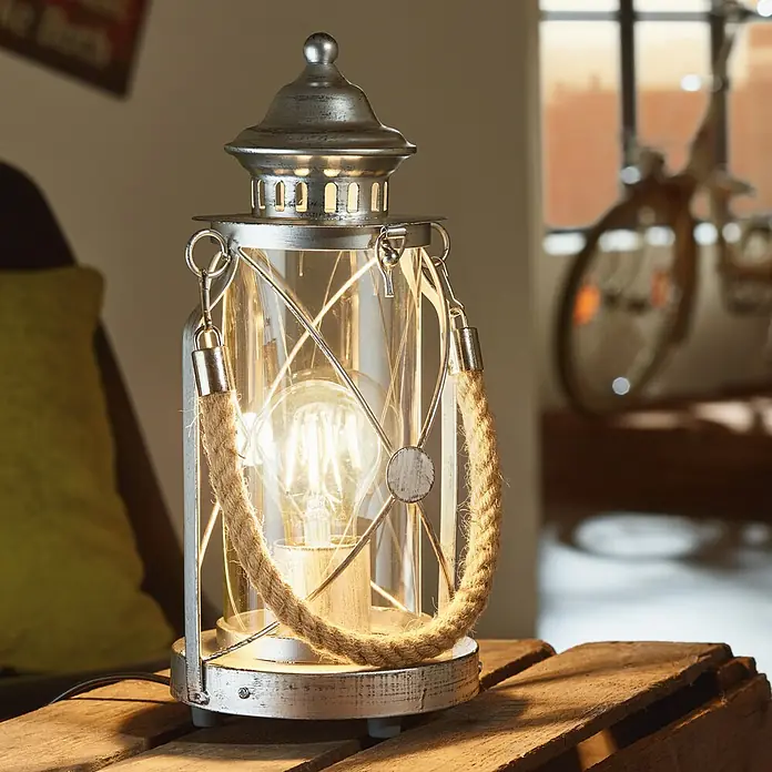 Eglo Tischleuchte Bradford - 60 W, Silber, Ø x H: 14 x 33 cm - Eine dekorative Tischleuchte im Laternen-Design mit Glühbirne, Seilgriffen und silberfarbener Oberfläche steht auf einem Holztisch.