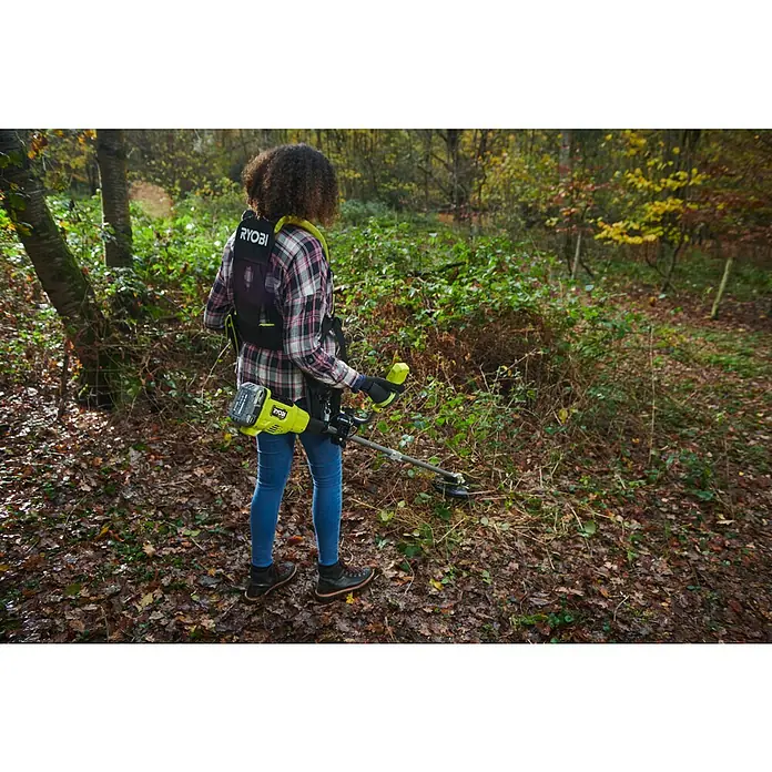 Ryobi ONE+HP Akku-Freischneider RY18BCX26A-0 - 18 V, Ohne Akku, Schnittbreite Faden: 38 cm, Schnittbreite Messer: 26 cm - Frau mit Ryobi Akku-Rasentrimmer im Wald. Das Gerät hat einen Tragegurt und Akku in Grün und Schwarz.