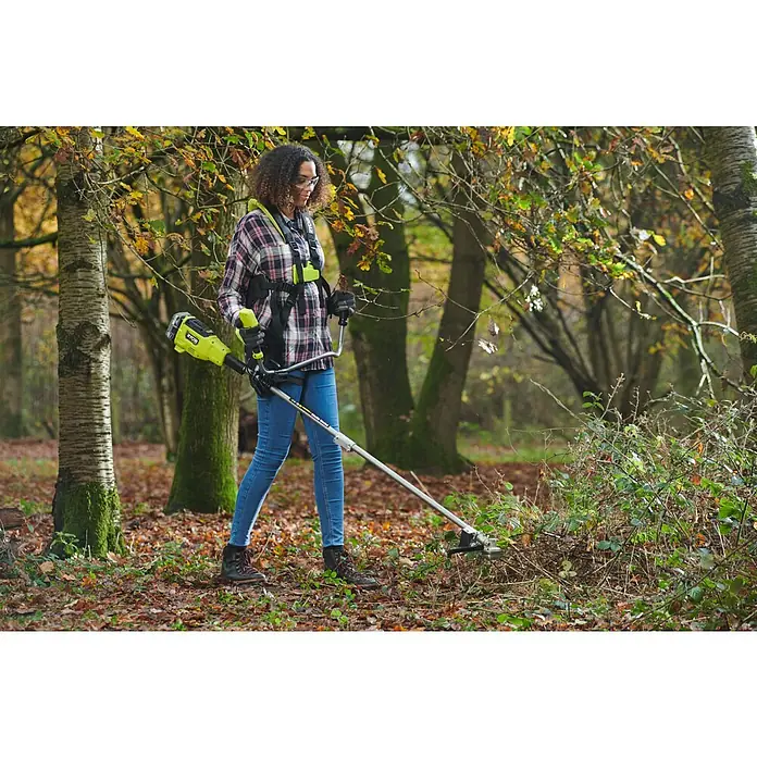Ryobi ONE+HP Akku-Freischneider RY18BCX26A-0 - 18 V, Ohne Akku, Schnittbreite Faden: 38 cm, Schnittbreite Messer: 26 cm - Eine Frau benutzt eine Ryobi-Akku-Motorsense, um Gestrüpp in einem herbstlichen Wald zu entfernen. Sie trägt Schutzkleidung und eine Tragevorrichtung.