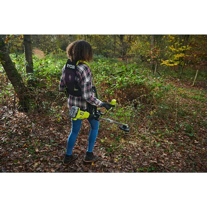 Ryobi ONE+HP Akku-Freischneider RY18BCX26A-0 - 18 V, Ohne Akku, Schnittbreite Faden: 38 cm, Schnittbreite Messer: 26 cm - Eine Person benutzt einen Ryobi-Rasentrimmer mit Akku und Rückentragegurt in einem Waldstück.