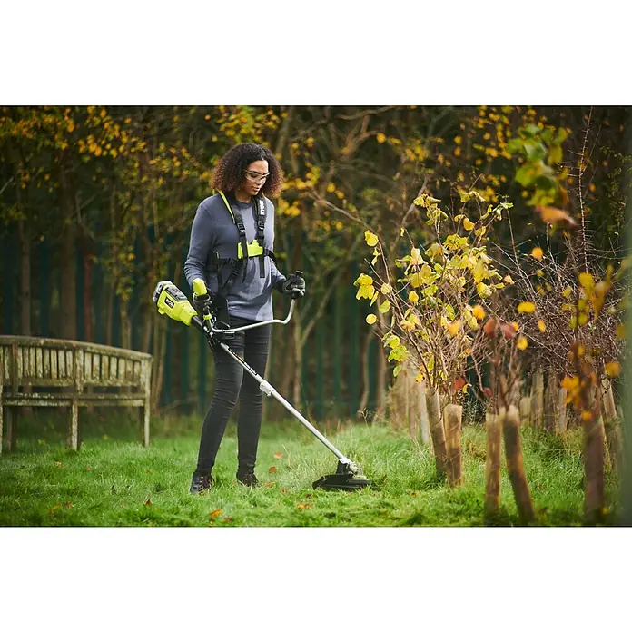 Ryobi ONE+HP Akku-Freischneider RY18BCX26A-0 - 18 V, Ohne Akku, Schnittbreite Faden: 38 cm, Schnittbreite Messer: 26 cm - Eine Frau, die einen Ryobi Rasentrimmer mit Gurt benutzt, um Gras in einem Garten mit Bäumen und einer Bank zu trimmen.