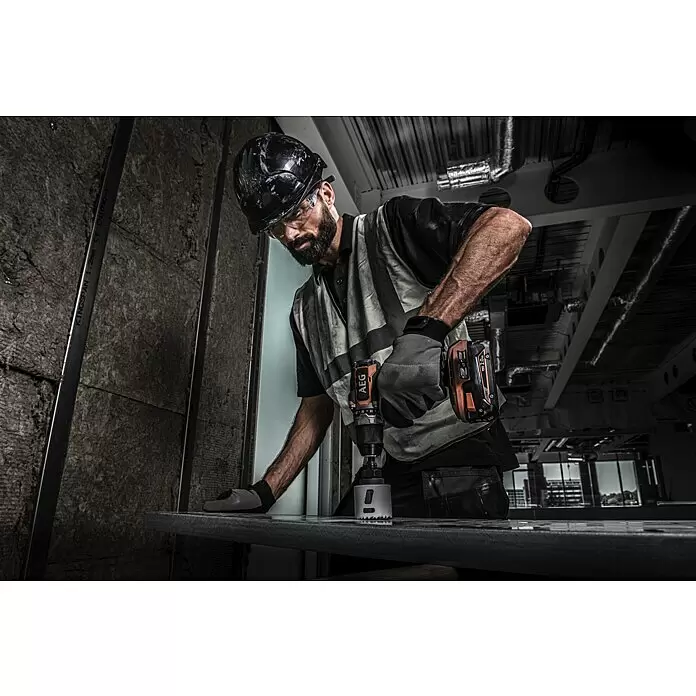 AEG Pro 18V Taladro atornillador de batería BS18SBL-202C - 18 V, 2 baterías, 2 Ah, 60 Nm - Un trabajador de la construcción usa un taladro AEG naranja y negro con una sierra de corona para cortar una superficie metálica.
