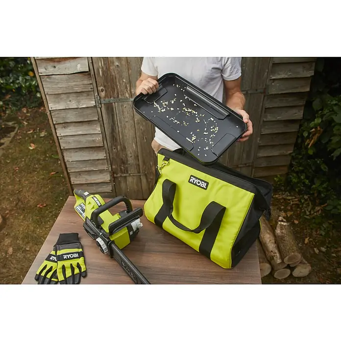 Ryobi Aufbewahrungstasche RAC256 - Passend für: 18 V und 36 V Ryobi Kettensägen - Ryobi Werkzeuge: Akku-Kettensäge, Tasche und Handschuhe auf einem Tisch. Im Hintergrund eine Person, die ein Tablett hält.