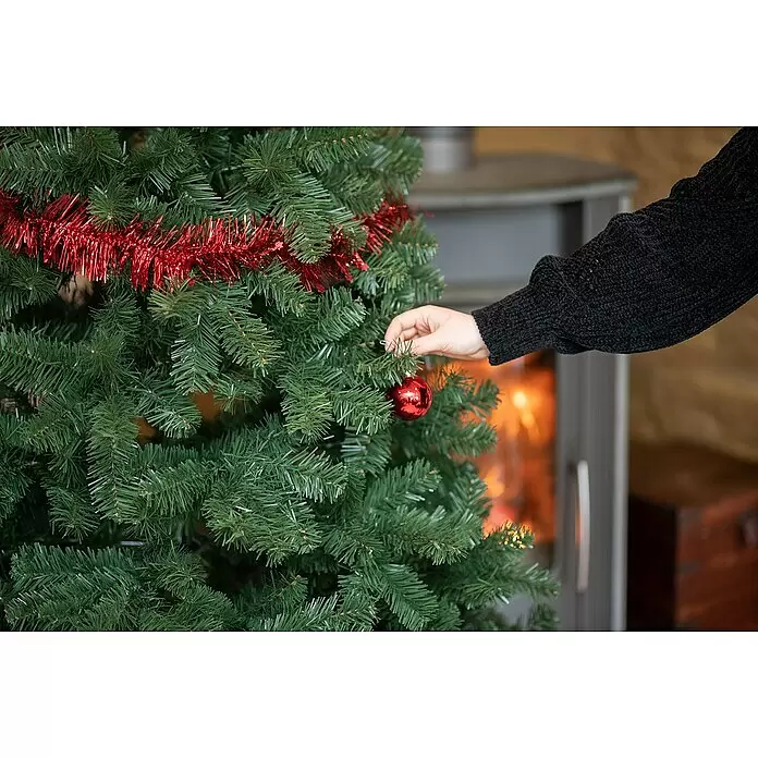 Künstlicher Weihnachtsbaum Oxford - Höhe: 210 cm - Eine Hand hängt eine glänzende rote Kugel an einen grünen Weihnachtsbaum, mit warmem Kamin im Hintergrund.
