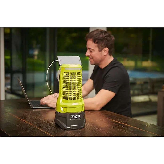 Ryobi ONE+ Insektenvernichter RY18BZA 0 - 18 V, Ohne Akku, Wirkungsbereich: 30 m² - Ein RYOBI-Gerät mit einem Handy oben drauf und einem Kabel, das es mit Strom versorgt, während eine Person an einem Laptop arbeitet.