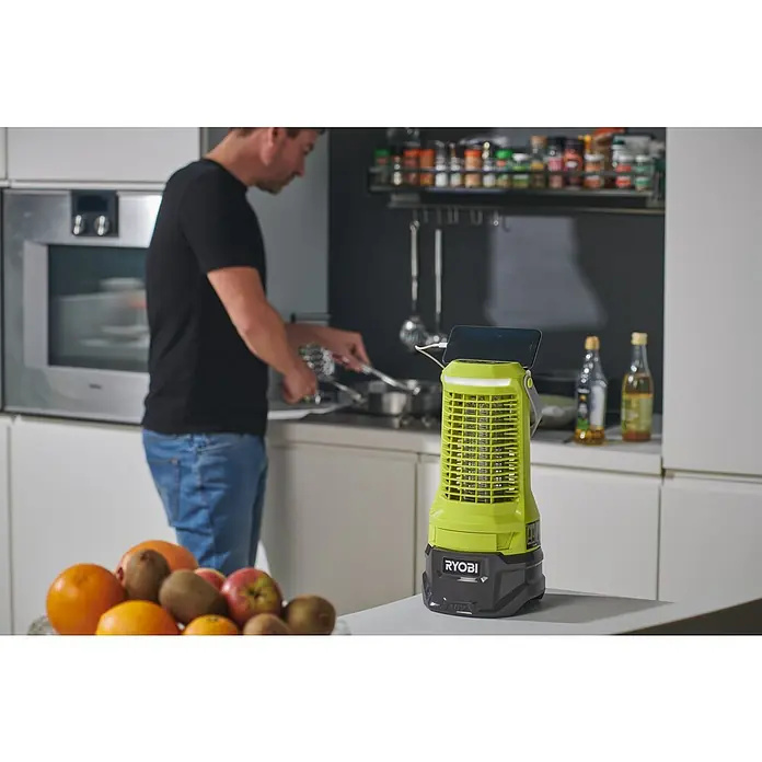 Ryobi ONE+ Insektenvernichter RY18BZA 0 - 18 V, Ohne Akku, Wirkungsbereich: 30 m² - Nahaufnahme eines Ryobi-Geräts zum Aufladen eines Mobiltelefons in einer modernen Küche, während eine Person im Hintergrund kocht.