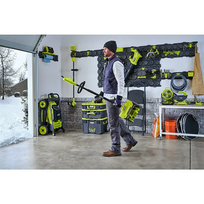 Ryobi ONE+ Akku-Schneefräse RY18ST25A-0 - 18 V, Ohne Akku, Arbeitsbreite: 25 cm - Ein Mann trägt Ryobi-Gartengeräte in einer Garage mit einem Ryobi-Wandsystem.